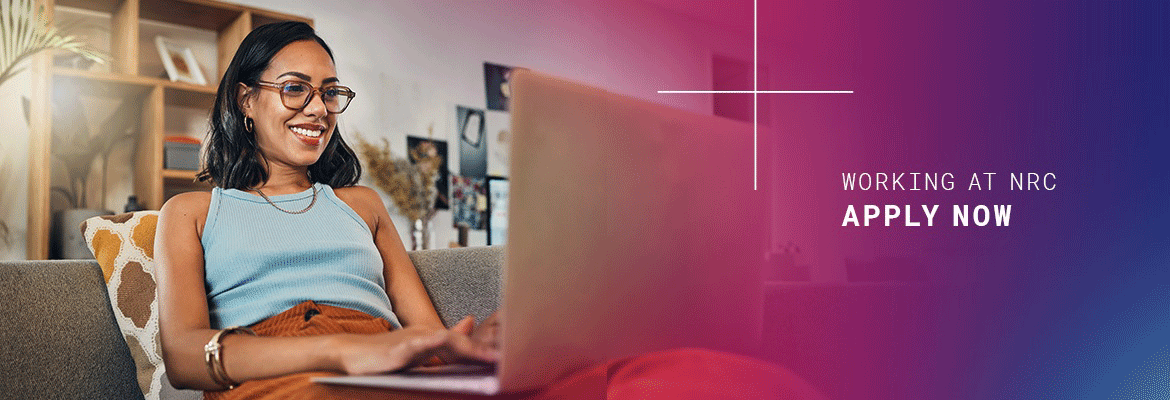 Black and white photo of a person sitting with their arms extended and typing on an opened laptop computer on a desk, with a drinking glass half full of water, and a cup of soup with lid on sitting in a small bowl with spoon along side, and a pair of folded reading glasses laying on the table next to the laptop, with the words 'working at nrc' and (in red font and larger text) 'apply now' in larger font and to the right of the photo on a white background.