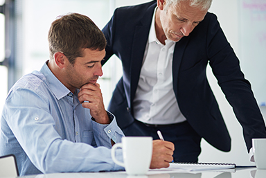 Two male budget analyst employees looking down at a document on a table.  One is seated holding a pen to the document with his left hand on his chin, and the other employee is standing, leaning over onto the table with his left arm