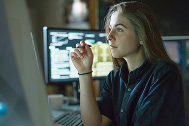 photo of a woman holding a pen in her hand with her elbow propped on a table, looking forward toward what you assume is a computer monitor. To her right in the background is another computer monitor.