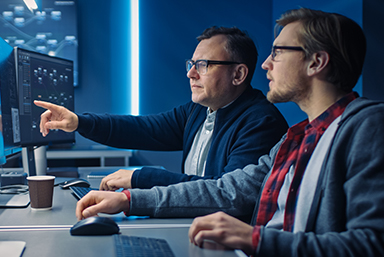 Two men seated at a computer table looking toward a monitor, which does not appear in the photo. 
 There is a monitor to the man on the right, who is pointing forward with his arm extended -- to what is probably a computer monitor.