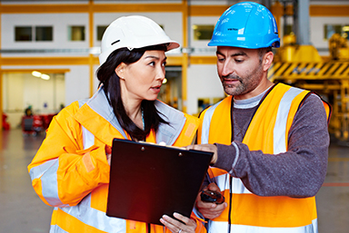 A female enforcement specialist, wearing a white hardhat and holding a clip board looking towards the worker next to her - a man with a blue hardhat and pointing at somethiing on the clip board.