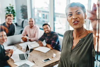 photo of a woman pointing at something on a board or display stand in a conference room, with four coworkers seated at a table behind her, looking at what she is presenting while smiling.