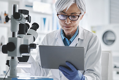 photo of a woman wearing a white lab coat standing in front of a large microscope and look down at a computer tablet she is holding in her left hand, with a latex glove.