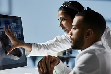 photo of a woman and man (seated) wearing white lab coats.  The woman is touching her hand to a computer monitor in front of them as they both look on.