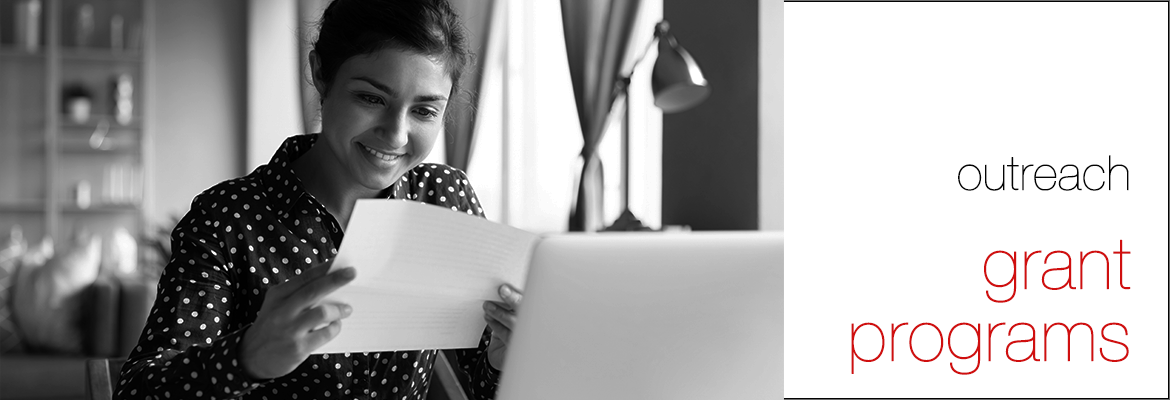 Black and white photo of a woman holding a sheet of paper in her hands reading it and smiling, with an open laptop in front of her, with the words 'outreach' and (in red font and larger text) 'grant programs' to the right of the photo on a white background.