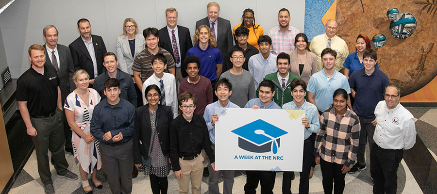 Picture of thirteen NRC adult employees and 18 high school students from diverse ethnic backgrounds. Three students in the center of the group are holding large sign with the 'A WEEK AT THE NRC' logo.