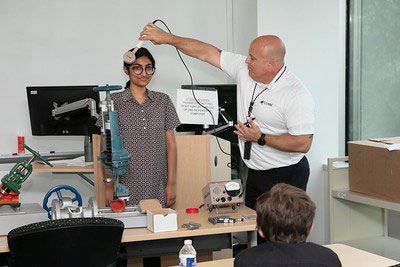 Picture of a classroom setting, in the front of the class a male instructor demonstrates the radioactive measurement tool on a female student, while other students look on from their seats.