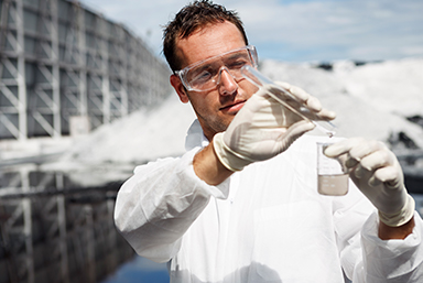 photo of a man with a white lab coat on wearing latex gloves, is holding a long test tube in his right hand, and pouring it into a clear glass container held in his left hand.