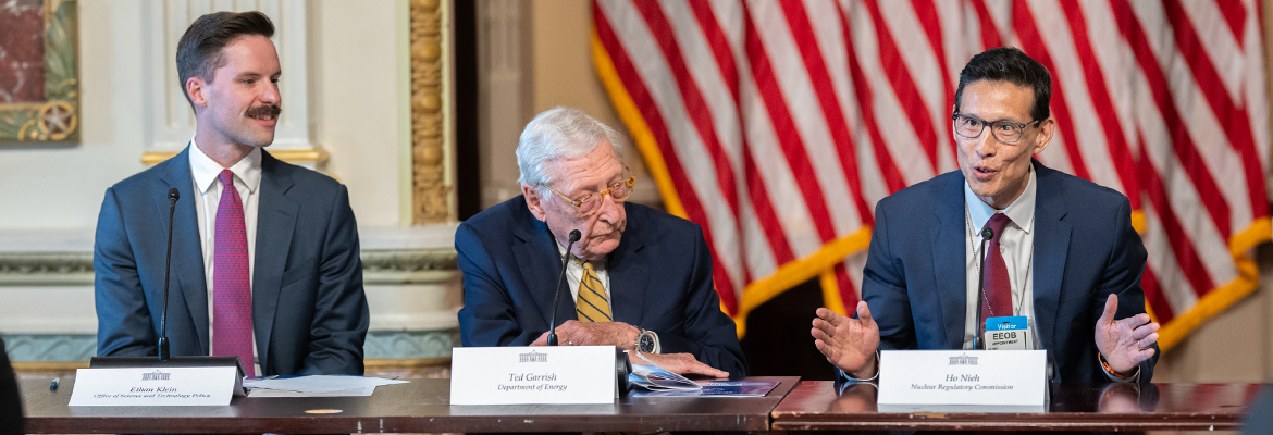 Three men sit at a long table speaking during a panel at the UPRISE (Utility Power Reactor Incremental Scaling Effort) event at the White House. The man on the right gestures with his hands while talking into a microphone, the older man in the center looks down at documents, and the man on the left watches with a slight smile. Name placards, microphones and papers are on the table, and a large American flag hangs in the background.
