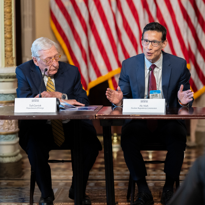 Two men sit at a long table speaking during a panel at the UPRISE (Utility Power Reactor Incremental Scaling Effort) event at the White House. The man on the right gestures with his hands while talking into a microphone, the older man in the center looks down at documents, and the man on the left watches with a slight smile. Name placards, microphones and papers are on the table, and a large American flag hangs in the background.