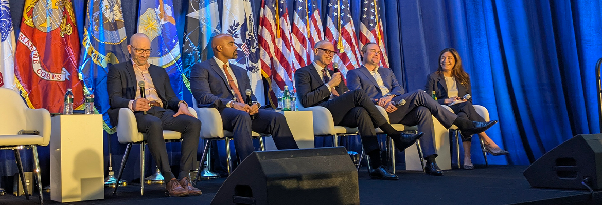 Image is a photo of five people sitting on a stage speaking on a panel.