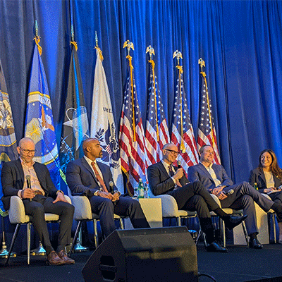 Image is a photo of five people sitting on a stage speaking on a panel.