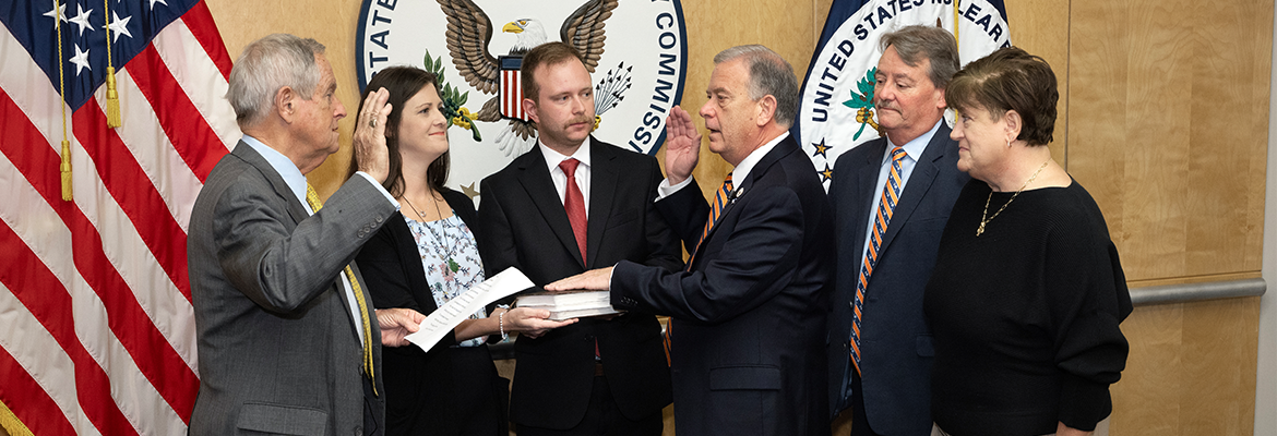 Image is a photo of six people standing. Two people have their hands up and one has his hand on a book. There is an American flag, NRC flag, and NRC seal on the wall behind them.