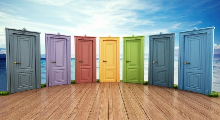 Seven closed wooden doors in a rainbow of colors standing in a curved row on a wooden deck.