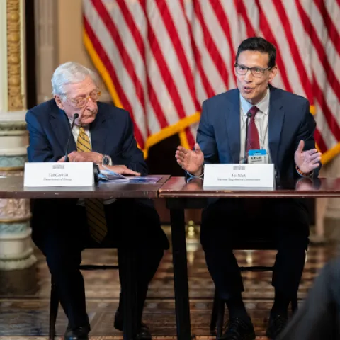 Two men sit at a long table speaking during a panel at the UPRISE (Utility Power Reactor Incremental Scaling Effort) event at the White House. The man on the right gestures with his hands while talking into a microphone, the older man in the center looks down at documents, and the man on the left watches with a slight smile. Name placards, microphones and papers are on the table, and a large American flag hangs in the background.
