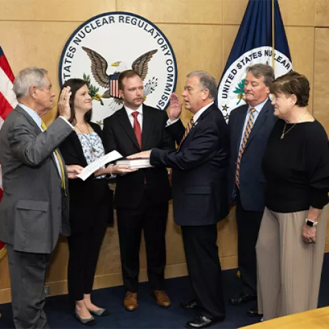 Image is a photo of six people standing. Two people have their hands up and one has his hand on a book. There is an American flag, NRC flag, and NRC seal on the wall behind them.
